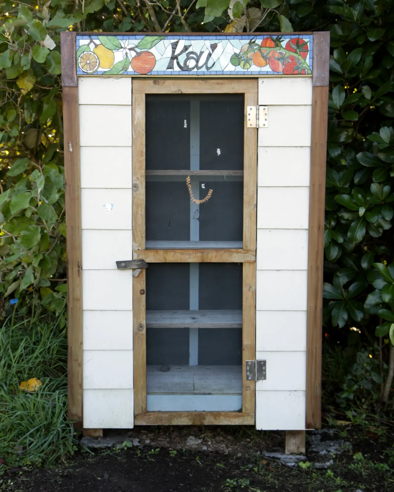 A small painted community food cupboard standing beside a garden hedge