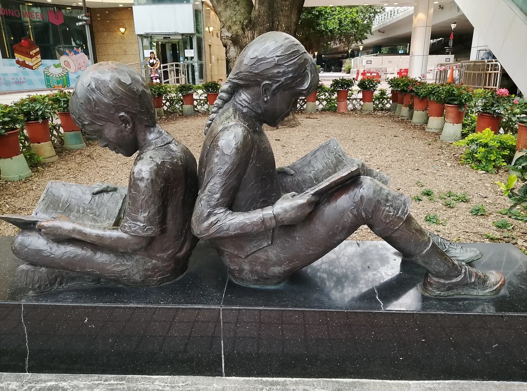 Bronze sculpture of two children sitting back to back reading books in a garden